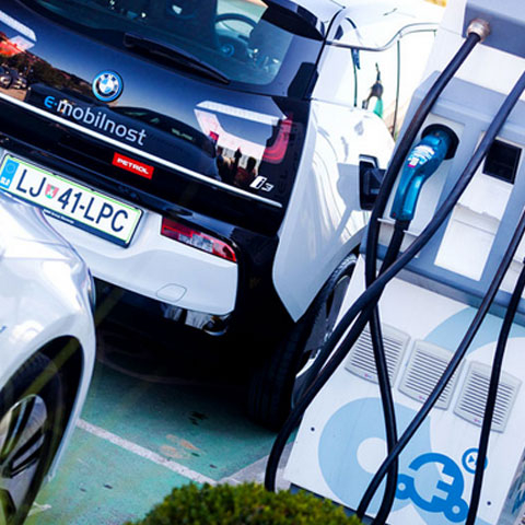 Na sliki sta dve električni vozili, od katerih eno, z oznako »e-mobilnost« in logotipom Petrol, polni baterijo na javni polnilnici. V ospredju je vidna bela polnilna postaja z modrim kablom in moder logo v obliki električnega priključka