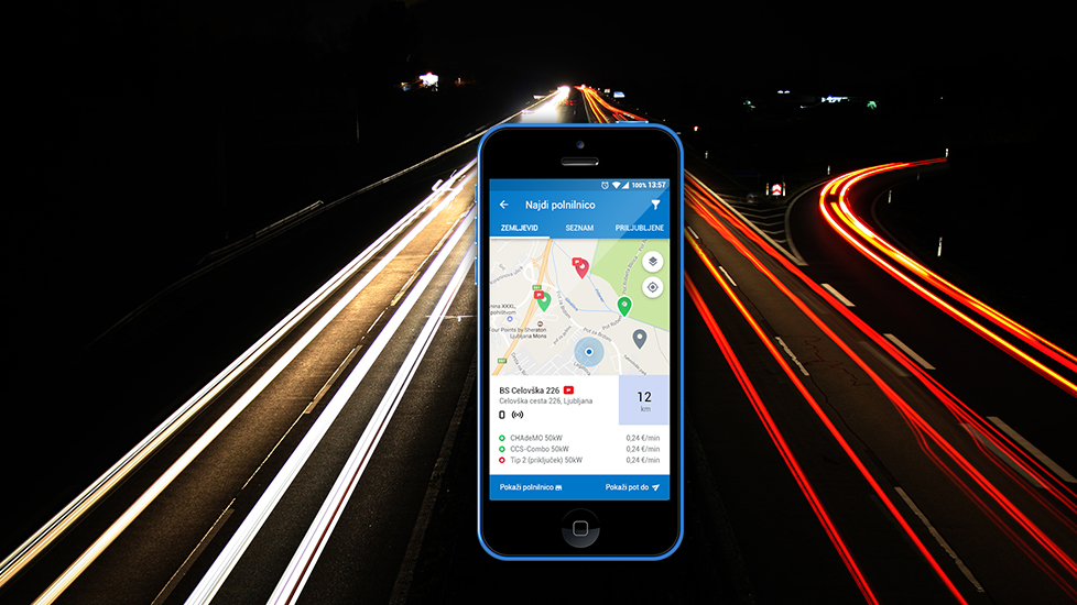 Na sliki je pametni telefon z odprto aplikacijo OneCharge za iskanje električnih polnilnic, prikazan na sredini nočne ceste z razpotegnjenimi svetlobnimi sledmi vozil v ozadju.