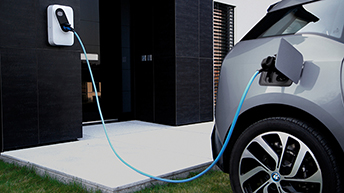 Na sliki je električni avtomobil, priključen na domačo stensko polnilnico z modrim kablom. Polnilnica je nameščena na temni fasadi sodobne hiše, vozilo pa je parkirano ob betonski ploščadi.