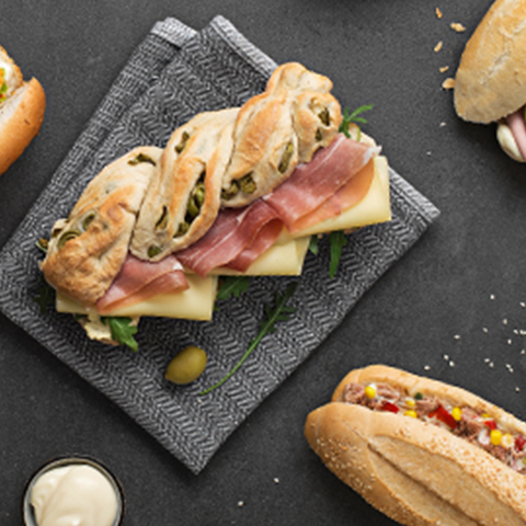 Na črnem krožniku so postreženi sveži sendviči z mesom, sirom in zelenjavo, poleg njih so še dodatni sendviči na svetlem ozadju.