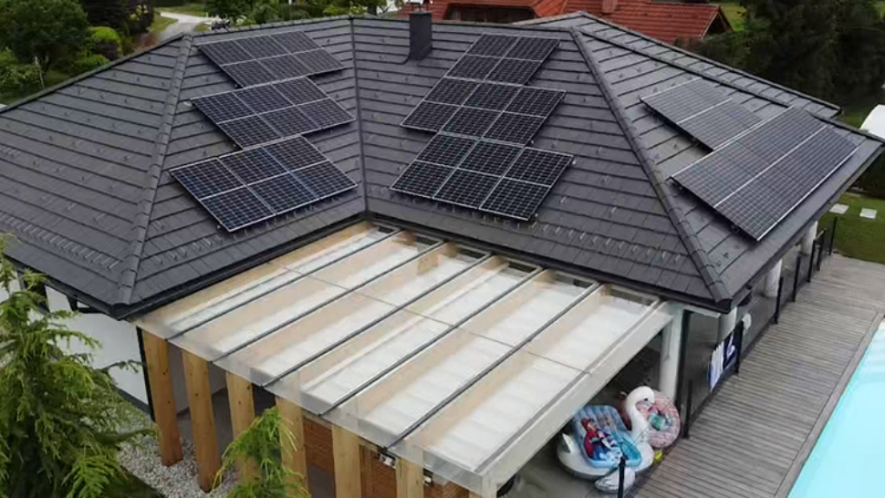 Na sliki je enonadstropna hiša s temno sivo streho, na kateri je nameščenih več nizov sončnih panelov. Panele vidimo na različnih straneh strehe, razporejene v pravokotnih sklopih. Pod streho je nadstrešek s prozorno streho iz stekla ali pleksi stekla, pod katerim stojijo leseni stebri. Ob hiši je lesena terasa, na kateri se nahaja bazen, ob njem pa nekaj napihljivih blazin in igrač. V ozadju so drevesa in sosednje hiše.