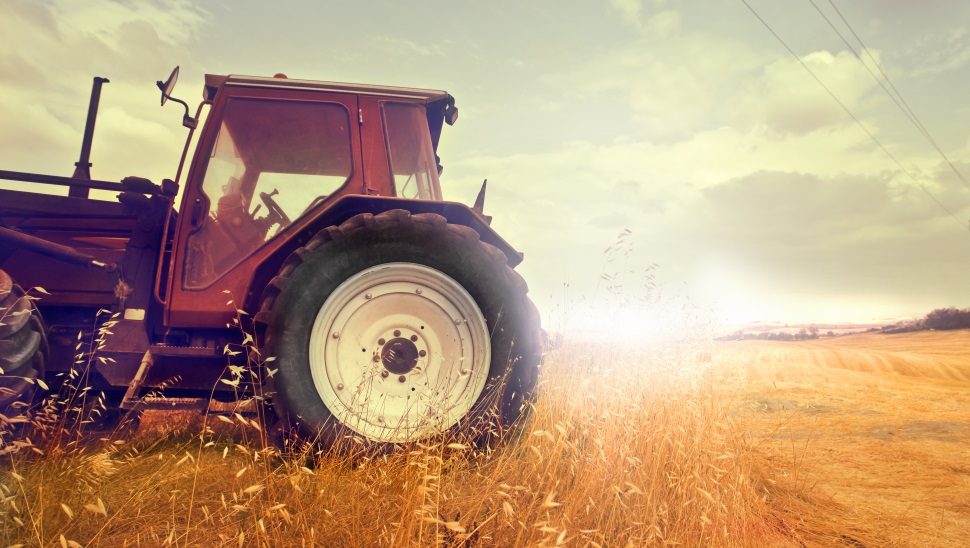 Na sliki je traktor, parkiran na robu polja, obdanega s travo in žitnimi rastlinami, z večernim sončnim zahodom v ozadju.