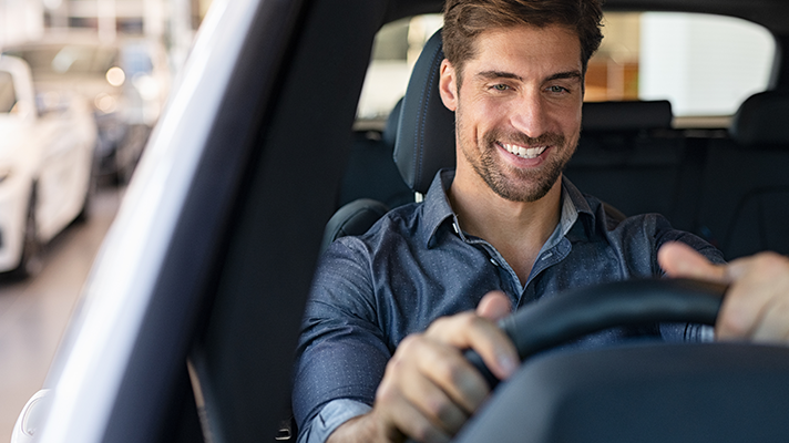 Na sliki je moški, ki sedi za volanom in se nasmejano vozi z avtomobilom. Videti je notranjost vozila iz voznikove perspektive.