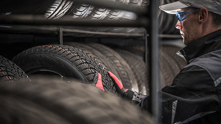 Na sliki je delavec v delovni obleki in rokavicah, ki pregleda oziroma namešča pnevmatiko na stojalu. Okoli njega so zložene avtomobilske gume.