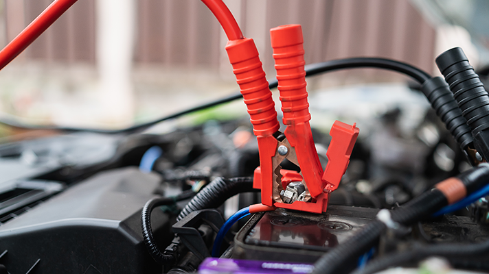 Na sliki je akumulator avtomobila, na katerega so priklopljene rdeče klešče za zagon motorja. Okoli so vidni kabli in drugi deli motornega prostora.