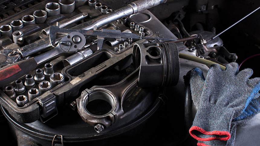 Na sliki je delovni pult z raztresenimi avtomehaničnimi orodji in delom motorja. Ob strani leži tudi delovna rokavica.