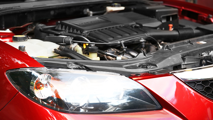 Na sliki je odprt pokrov rdečega avtomobila, skozi katerega je viden motorni prostor.