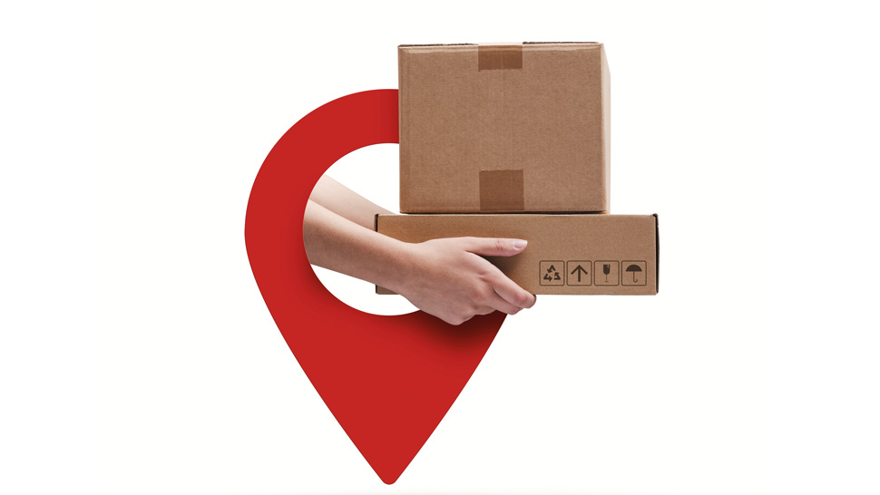 Roka drži dva kartonska paketa pred rdečo lokacijsko oznako na belem ozadju.