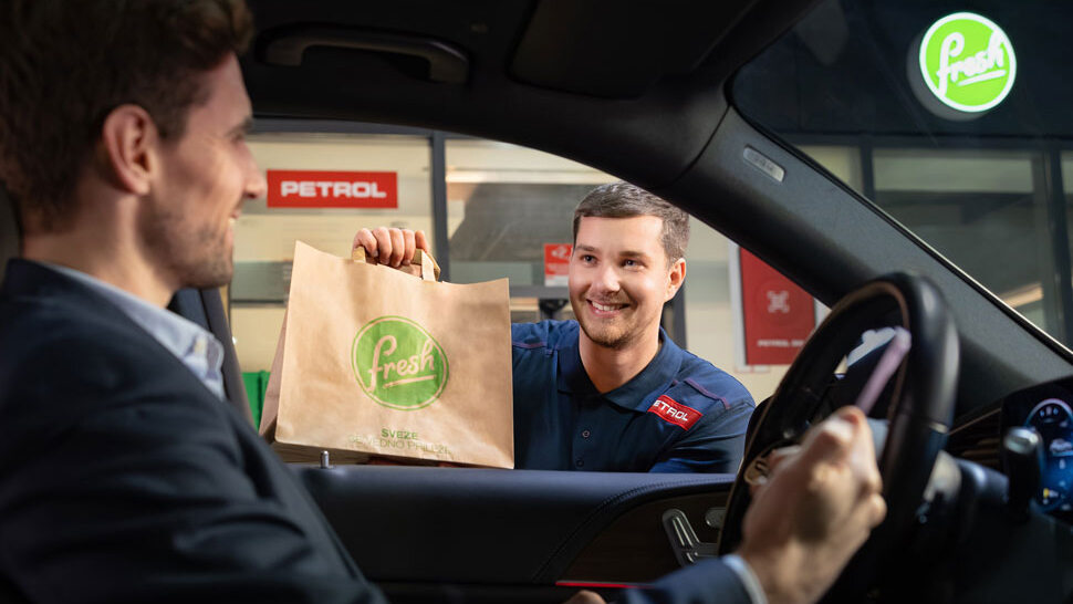 Na sliki prodajalec v Petrolovi uniformi, ki skozi okno avtomobila izroča papirnato vrečko z logotipom Fresh nasmejanemu vozniku v poslovni obleki. V ozadju sta vidna logotipa Petrol in Fresh na stavbi.