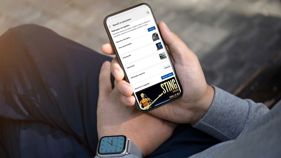 Na sliki oseba v sivi jopici in z uro na zapestju drži pametni telefon. Na zaslonu je odprta aplikacija z možnostjo nakupa vstopnic za dogodke, na dnu pa je prikazana reklama za koncert Stinga.
