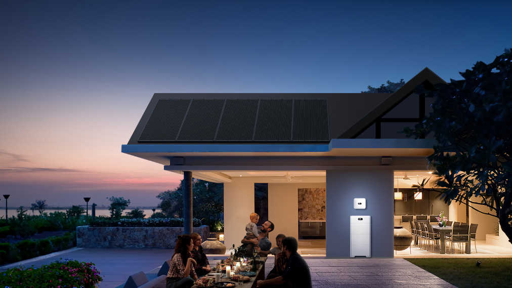 Na sliki je sodobna hiša v večernem času, osvetljena z umetno svetlobo iz notranjosti prostorov. Na strehi so nameščeni sončni paneli. Na fasadi hiše je vidna bela naprava, ki deluje kot hranilnik energije ali inverter za sončno elektrarno.