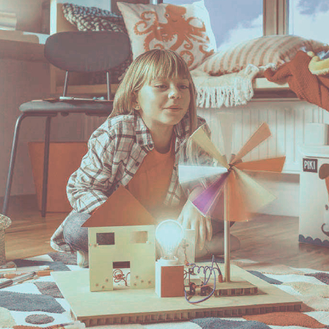 Na sliki je otrok, ki sedi na tleh v sobi in se igra z majhnim modelom hiše. Pred njim je maketa, ki prikazuje hišo, ob kateri stoji vetrnica z barvnimi kraki, priklopljena na preprosto električno vezje. Na vezju sveti žarnica.