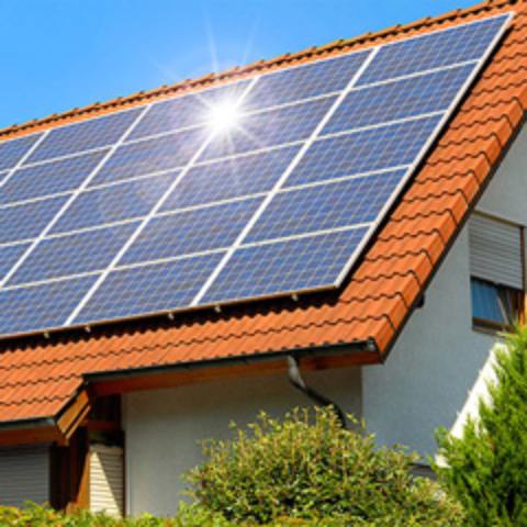 Na sliki je hiša z rdečo streho, na kateri so nameščeni sončni paneli. Plošče prekrivajo skoraj celotno površino strehe in so postavljene tako, da kar najbolje izkoriščajo sončno svetlobo. Sonce se močno odbija od ene izmed plošč.