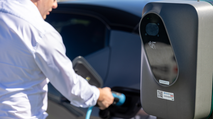 Na sliki je oseba, ki priklaplja električno vozilo na polnilnico podjetja Petrol. Polnilnica je v ospredju slike, medtem ko sta oseba in vozilo rahlo zamegljena v ozadju.