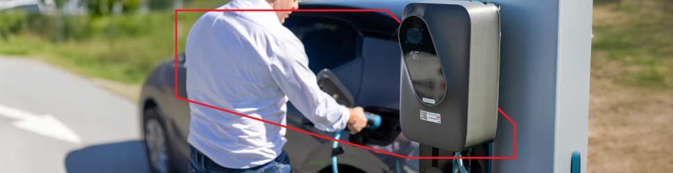 Na sliki je oseba, ki priklaplja električno vozilo na polnilnico podjetja Petrol, postavljeno ob cesti. V ozadju je naravno okolje, po celotni širini slike pa poteka rdeča geometrijska linija.