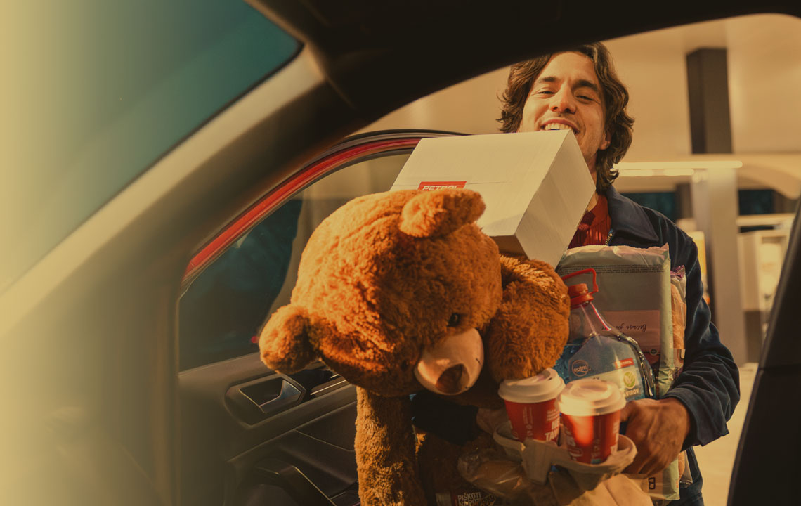 Oseba z nasmehom v rokah nosi veliko plišasto medvedko, dve kavi v lončkih, škatlo in več nakupovalnih vrečk, medtem ko stoji ob odprtih vratih avtomobila.