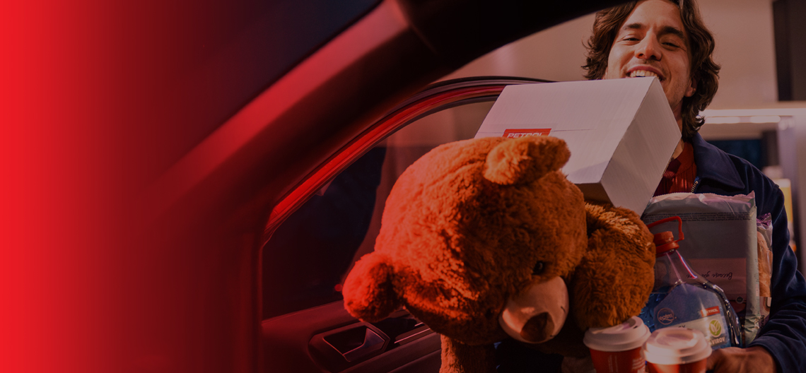 Na sliki je prikazana oseba, ki stoji ob odprtih vratih avtomobila in v rokah nosi več predmetov – velik plišast medved, škatla, dve vroči pijači v nosilcu ter plastenka pijače. Oseba se nasmejano nagiba proti vozilu, prizor pa je rahlo zatemnjen z rdečim prelivom na levi strani.