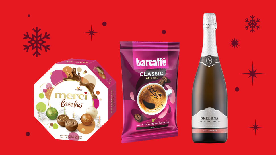 Na sliki je prikazano praznično darilno okolje z izbranimi živili in pijačo na rdečem ozadju, okrašenem s snežinkami. Na levi strani je škatla bonbonov Merci Lovelies v večbarvni embalaži. Na sredini je vrečka mlete kave Barcaffè Classic v rožnato-vijolični barvi z ilustracijo skodelice kave. Na desni strani stoji steklenica penečega vina Srebrna penina z belo etiketo in bleščečim srebrnim vratom. Slika deluje praznično in predstavlja predloge za darila.