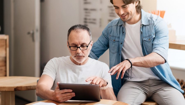 Na sliki je prikazan starejši moški, ki sedi za mizo in uporablja tablico, ob njem pa stoji mlajši moški, ki mu pomaga in mu svetuje, kar ponazarja medgeneracijsko sodelovanje, prenos znanja in podporo pri uporabi digitalnih tehnologij.