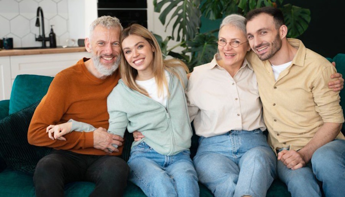 Na sliki je prikazana štiričlanska družina, ki sproščeno sedi skupaj na kavču v domačem okolju, objeta in nasmejana, kar ponazarja medgeneracijsko povezanost, toplino in občutek varnosti.