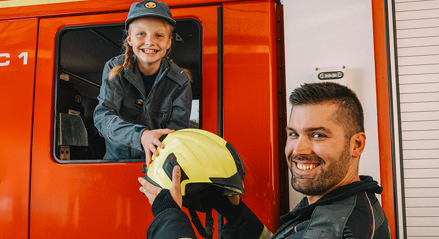 Na sliki je prikazana mlada deklica v gasilski uniformi, ki sedi v kabini gasilskega vozila in se nasmejano nagiba skozi odprto okno, medtem ko ji gasilec pred vozilom z nasmehom poda rumeno gasilsko čelado. Ozadje sestavlja rdeče gasilsko vozilo z vidnimi vrati in delom kabine.