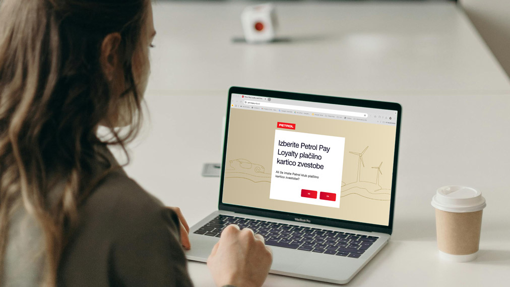Na sliki je oseba, ki sedi za mizo in uporablja prenosni računalnik, na katerem je odprta spletna stran z obrazcem za pridobitev plačilne kartice Petrol Pay Loyalty. Na zaslonu je prikazano spletno mesto z napisom »Izberite Petrol Pay Loyalty plačilno kartico zvestobe« ter gumbi za nadaljevanje postopka. V ozadju je mehko zamegljeno okolje sodobne pisarne z belo skodelico za kavo na mizi. Slika ponazarja izbiro in naročilo nove Petrol Pay Loyalty plačilne kartice zvestobe prek spletne strani.