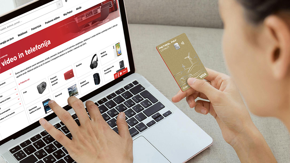 Na sliki je prikazano osebo, ki opravlja spletni nakup preko prenosnega računalnika in hkrati v roki drži plačilno kartico Petrol Pay Visa. Na zaslonu računalnika je odprta spletna trgovina Petrol, v kateri so prikazani različni izdelki iz kategorije avdio in video ter telefonija.