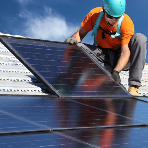 Na sliki je delavec s čelado in varnostnim pasom, ki namešča sončne panele na streho.