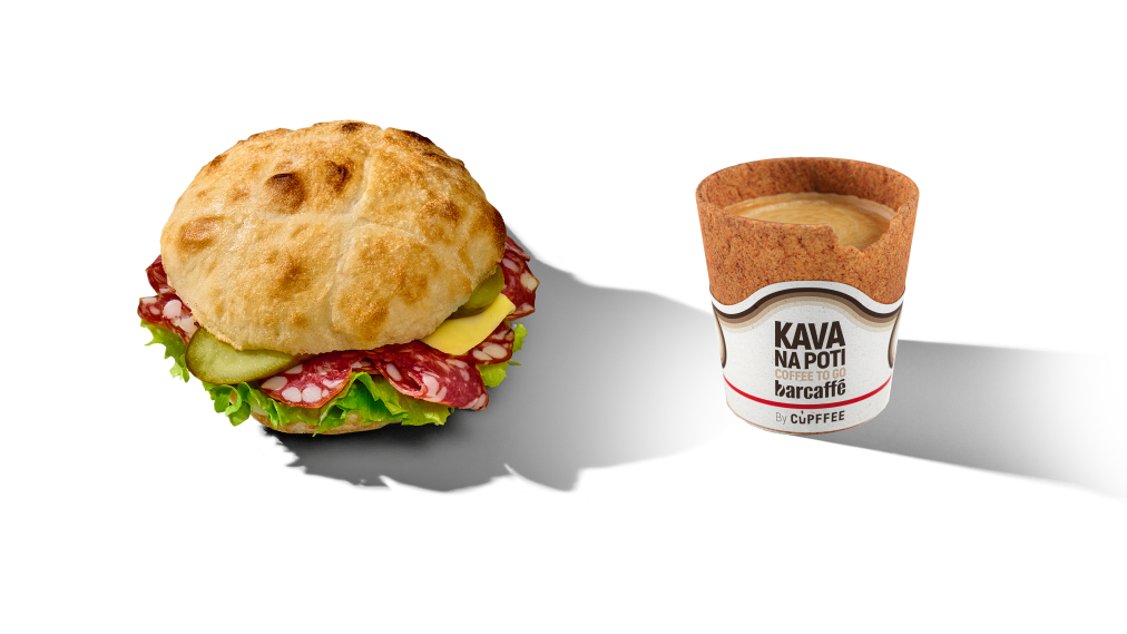 Sendvič s suho salamo, sirom, kislimi kumaricami in listom solate v hrustljavi okrogli bombetki poleg kave Na poti v užitnem kozarčku s potiskom 'Kava na poti – barcaffè by Cupffee'.