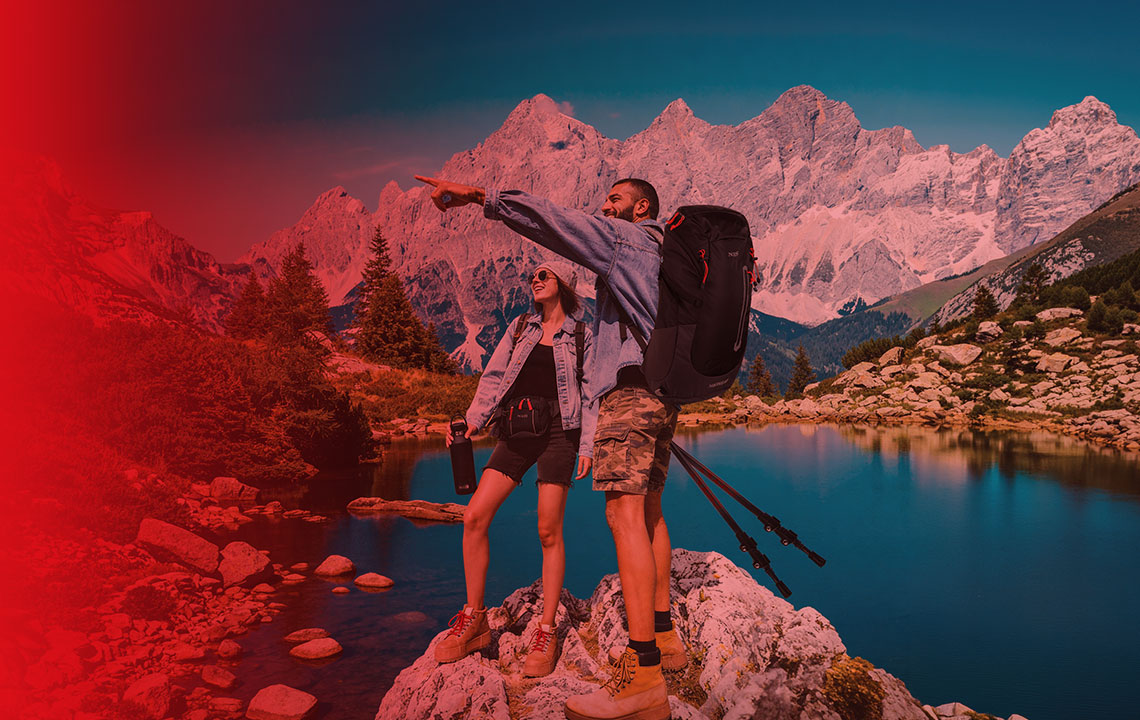 Dve osebi stojita ob alpskem jezeru s pohodniško opremo; ena oseba drži pohodne palice, druga steklenico, v ozadju pa se dvigajo visoke skalnate gore.