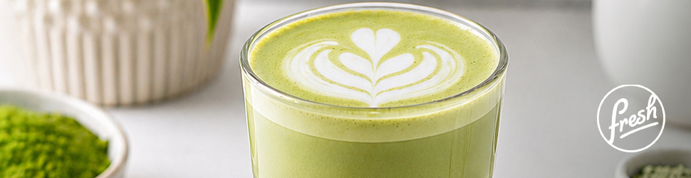 Kozarec svetlo zelene matcha latte pijače z mlečno peno in dekorativnim vzorcem v obliki srca na vrhu. V ozadju so bela posoda, skodelica z zelenim prahom matche in rastlina, kar ustvarja čist, minimalističen ambient. Desno spodaj je logo Fresh.