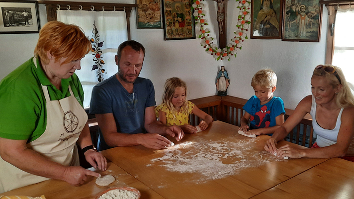 Na sliki je prikazana družina skupaj z starejšo žensko pri pripravi testa za peko za leseno mizo v tradicionalno opremljenem prostoru. Odrasli in otroci sodelujejo pri oblikovanju testa, vzdušje pa je domače in sproščeno.