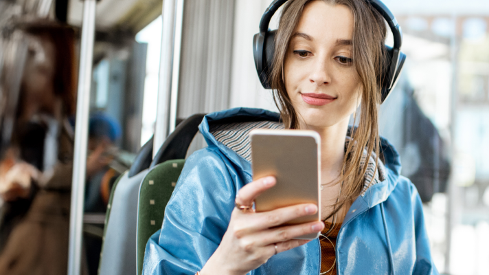 Na sliki je oseba s slušalkami, ki med vožnjo z javnim prevozom na mobilnem telefonu uporablja aplikacijo za poslušanje zvočnih knjig. Prizor ponazarja dostop do digitalnih vsebin, branja in poslušanja knjig kjerkoli in kadarkoli.