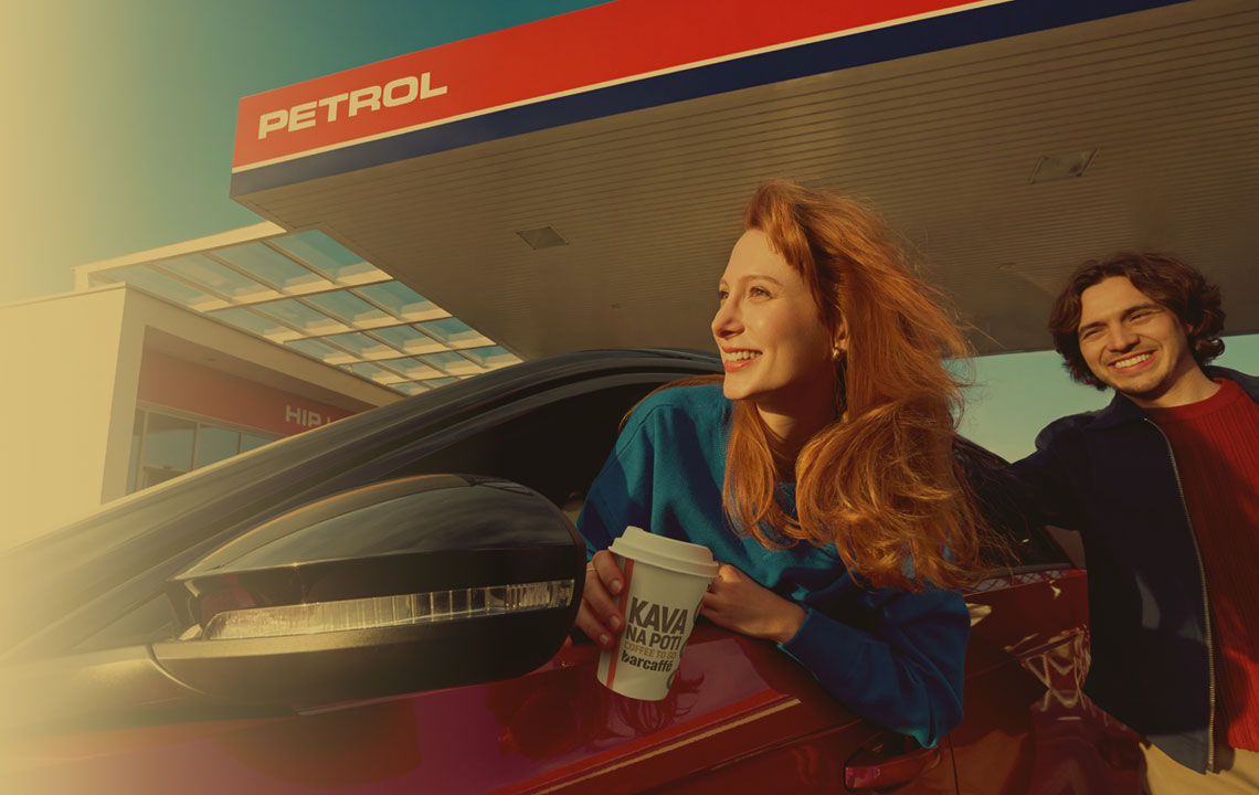 Nasmejana mlada ženska z lončkom kave sedi v avtomobilu na bencinskem servisu Petrol, medtem ko ob njej stoji mlad moški. Prizor simbolizira prijetno uporabniško izkušnjo, postanek na poti in sproščeno mobilnost.