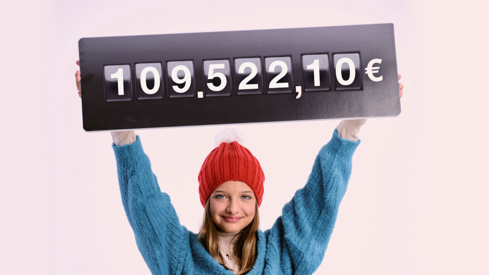 Na sliki je prikazana deklica z rdečo kapo, ki z dvignjenimi rokami nad glavo drži tablo z zapisanim zneskom 109522,10 €.