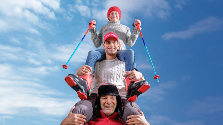 Na sliki so tri osebe, postavljene ena na drugo na prostem pod modrim nebom z oblaki. Spodaj je nasmejana oseba v rdeči jakni in črni zimski kapi. Na njenih ramenih sedi mlajša oseba v svetlem pletenem puloverju in rdeči kapi. Na vrhu sedi še ena oseba, oblečena v siv pulover, modre kavbojke in rdečo kapo, ki v rokah drži smučarske palice. Vse tri osebe delujejo razigrano in nasmejano.