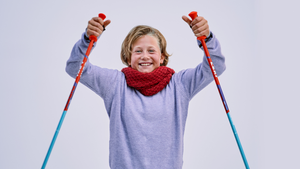 Na sliki je nasmejana oseba s kratkimi sivimi lasmi, oblečena v svetlo vijoličen pulover in rdeč šal. V rokah drži palice za nordijsko hojo, dvignjene navzgor, kot da telovadi ali praznuje. Ozadje je svetlo in nevtralno.