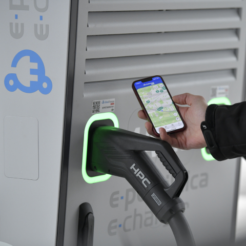 Na sliki je prikazana električna polnilnica za vozila, v katero je priključen polnilni kabel. Oseba v roki drži pametni telefon z odprto aplikacijo za upravljanje ali iskanje polnilnic. Okoli priključkov na polnilnici svetijo zeleni LED-obroči, v ozadju pa so vidni grafični elementi, povezani z električno mobilnostjo in e-polnjenjem.