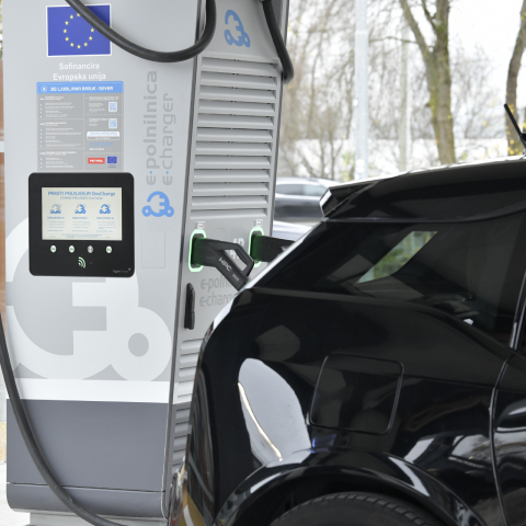 Na sliki je prikazano električno vozilo, priključeno na javno električno polnilnico. V ospredju je vidna polnilna postaja z zaslonom, navodili za uporabo in priključenim polnilnim kablom, ki je povezan z vozilom. Prizor ponazarja polnjenje električnega vozila na javni polnilni infrastrukturi ter uporabo storitev e-mobilnosti.