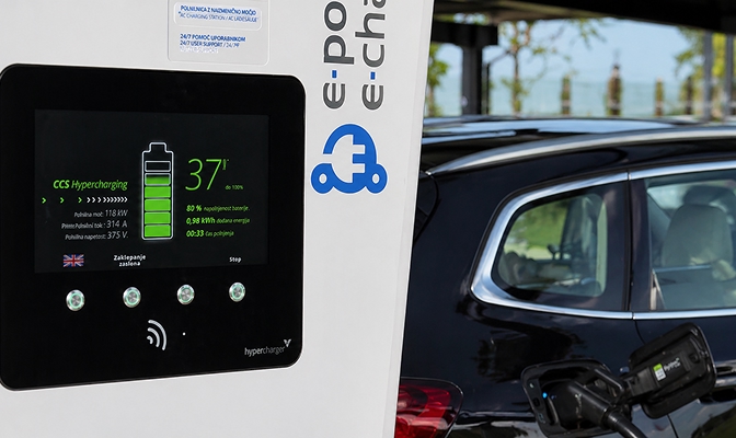 Na sliki je prikazana hitra polnilna postaja za električna vozila med postopkom polnjenja. Na zaslonu polnilnice so vidni podatki o stanju baterije in poteku polnjenja, ob njej pa stoji električni avtomobil, priključen na polnilni kabel. Fotografija poudarja uporabo infrastrukture za e-mobilnost.