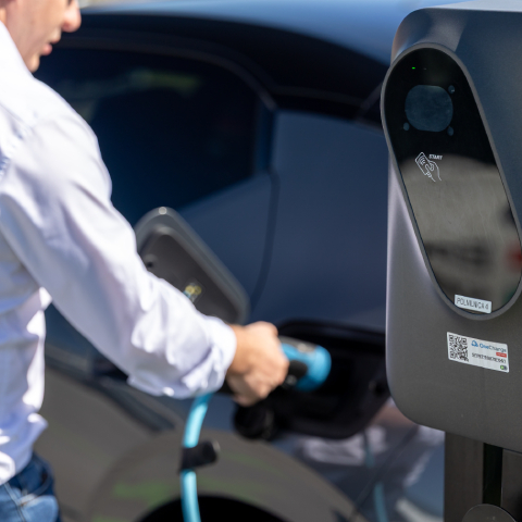 Na sliki je prikazano električno vozilo med polnjenjem na javni električni polnilnici. Oseba priključuje polnilni kabel v vozilo, v ospredju pa je vidna polnilna postaja z brezstičnim označevalnikom za identifikacijo ali plačilo. Prizor ponazarja uporabo rešitev za polnjenje električnih vozil in trajnostno mobilnost.