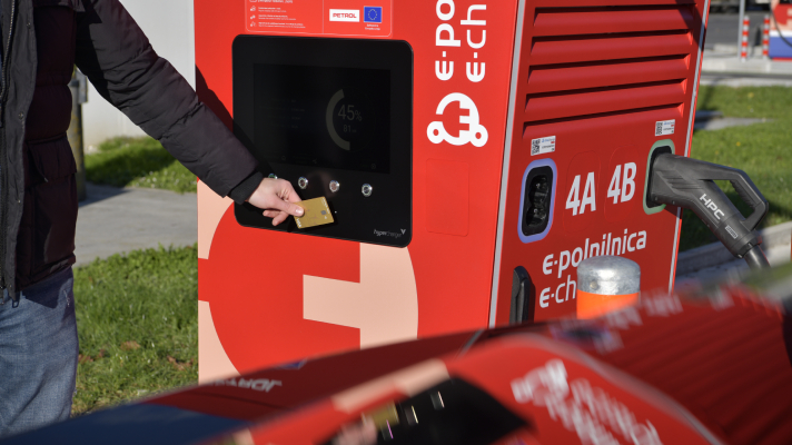 Na sliki je polnilna postaja za električna vozila rdeče barve. Oseba pred zaslonom polnilnice drži kartico, ob strani pa je priključen polnilni kabel. Na polnilnici so vidni napisi »e-polnilnica«, »e-charge« ter oznaki priključkov »4A« in »4B«.
