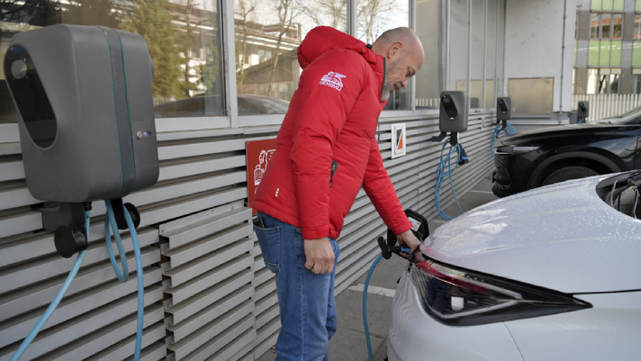 Na sliki je oseba, ki priključuje električno vozilo na polnilnico ob stavbi, v ozadju pa so vidne dodatne polnilne postaje.