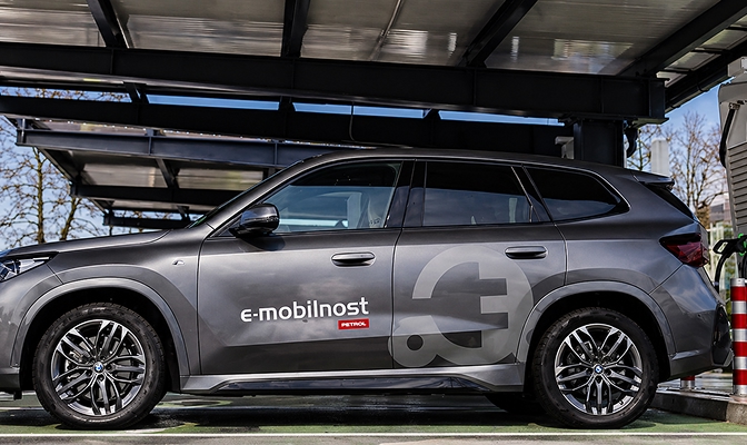 Siv električni SUV z napisom »e-mobilnost« je parkiran ob polnilnici za električna vozila pod nadstreškom. Vozilo je prikazano s strani, v ozadju pa so vidni polnilna infrastruktura in drevesa.