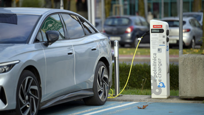 Na sliki je električni avtomobil, priključen na polnilno postajo za električna vozila. Ob vozilu stoji bela polnilnica z napisom »e-polnilnica« in »e-chargen«, v katero je priključen polnilni kabel. V ozadju so vidna parkirana vozila in poslovna stavba.