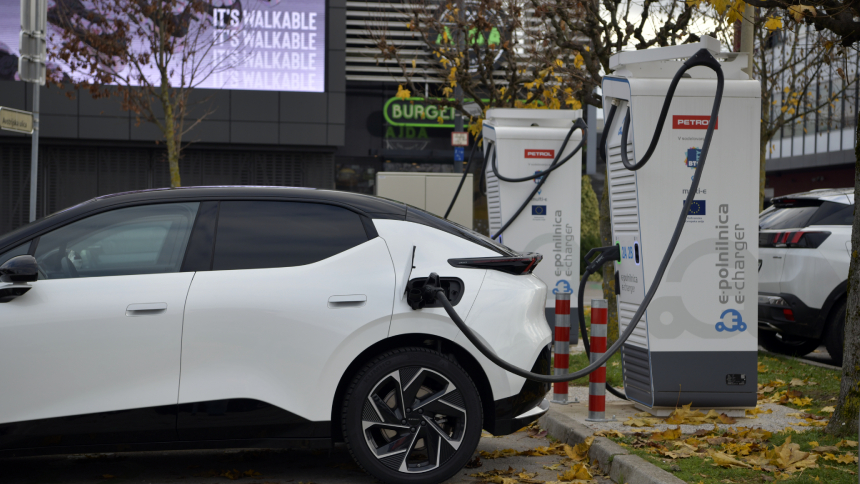 Na sliki je belo električno vozilo, priključeno na polnilno postajo za električna vozila. Polnilni kabel je povezan med vozilom in polnilnico z napisom »e-polnilnica e-charge«. V ozadju so vidne dodatne polnilne postaje, parkirana vozila, drevesa in poslovne stavbe.