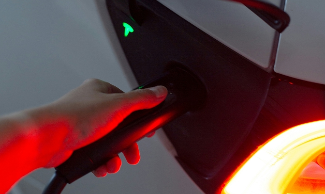 Na sliki je prikazan postopek polnjenja električnega vozila. Oseba z roko drži polnilni priključek, ki je vstavljen v polnilno vtičnico avtomobila. Vidni so svetlobni indikatorji in osvetljen zadnji del vozila, ki nakazujejo aktivno polnjenje.