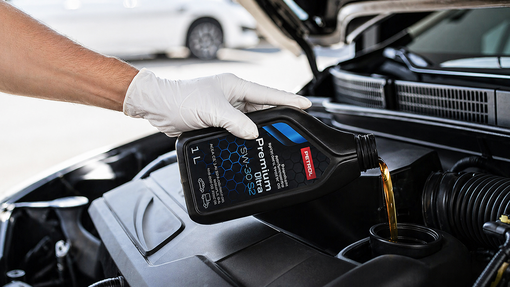 Roka v beli zaščitni rokavici doliva motorno olje Petrol Premium Ultra v motorni prostor avtomobila. Pokrov motorja je odprt, olje pa teče iz črne plastenke v odprtino za dolivanje.