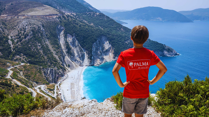 Otrok v rdeči majici z napisom Palma stoji na razgledni točki nad turkiznim zalivom in belo prodnato plažo, obdanima z visokimi skalnatimi pečinami ter modrim morjem.