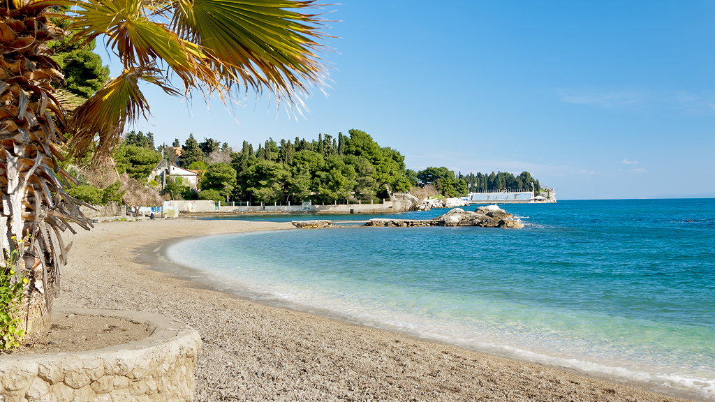 Mirna prodnata plaža ob turkiznem morju, obdana z borovci in sredozemskim rastlinjem. V ospredju so palma in kamnit zid, v ozadju pa obala z belimi stavbami pod jasnim modrim nebom.